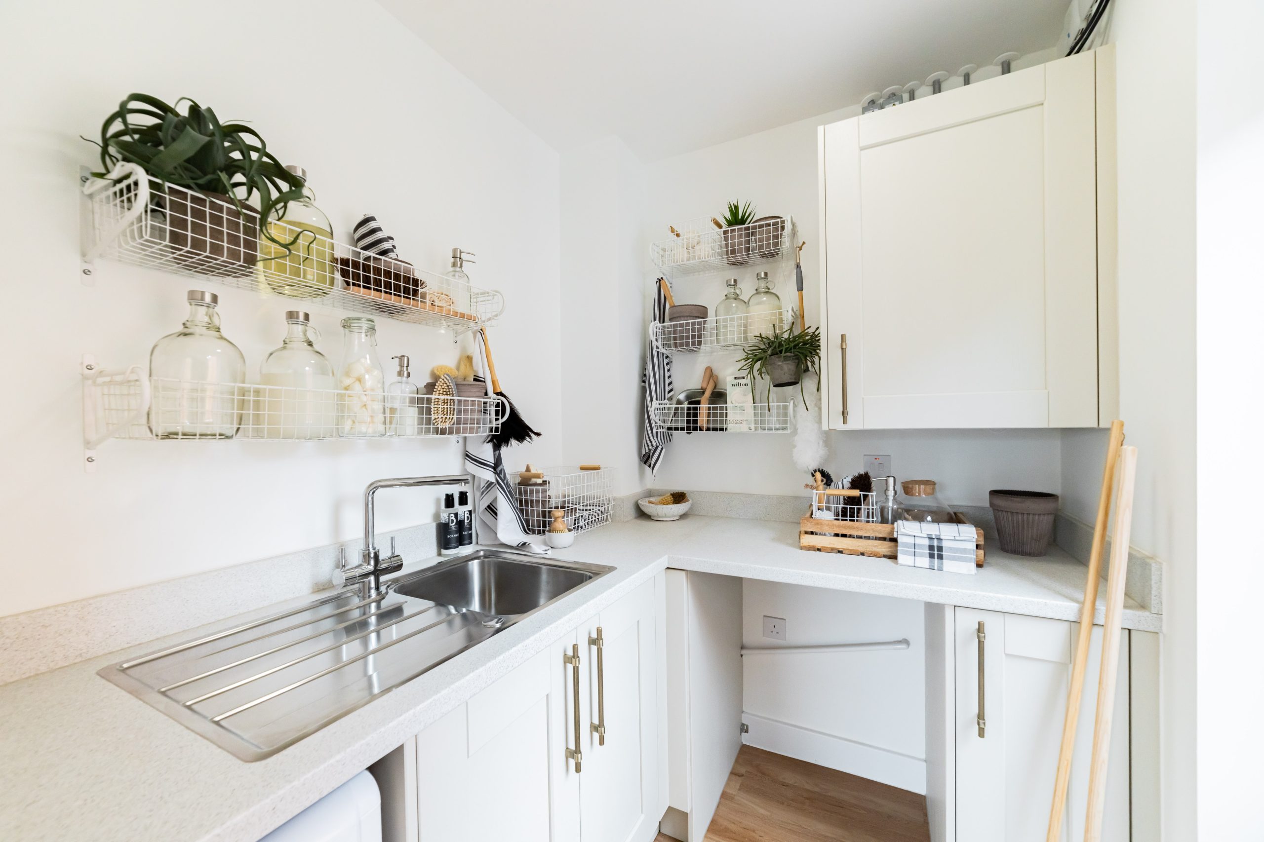 Bright utility room with stainless steel sink, white shaker cabinets, and open wire shelving displaying glass jars and potted plants