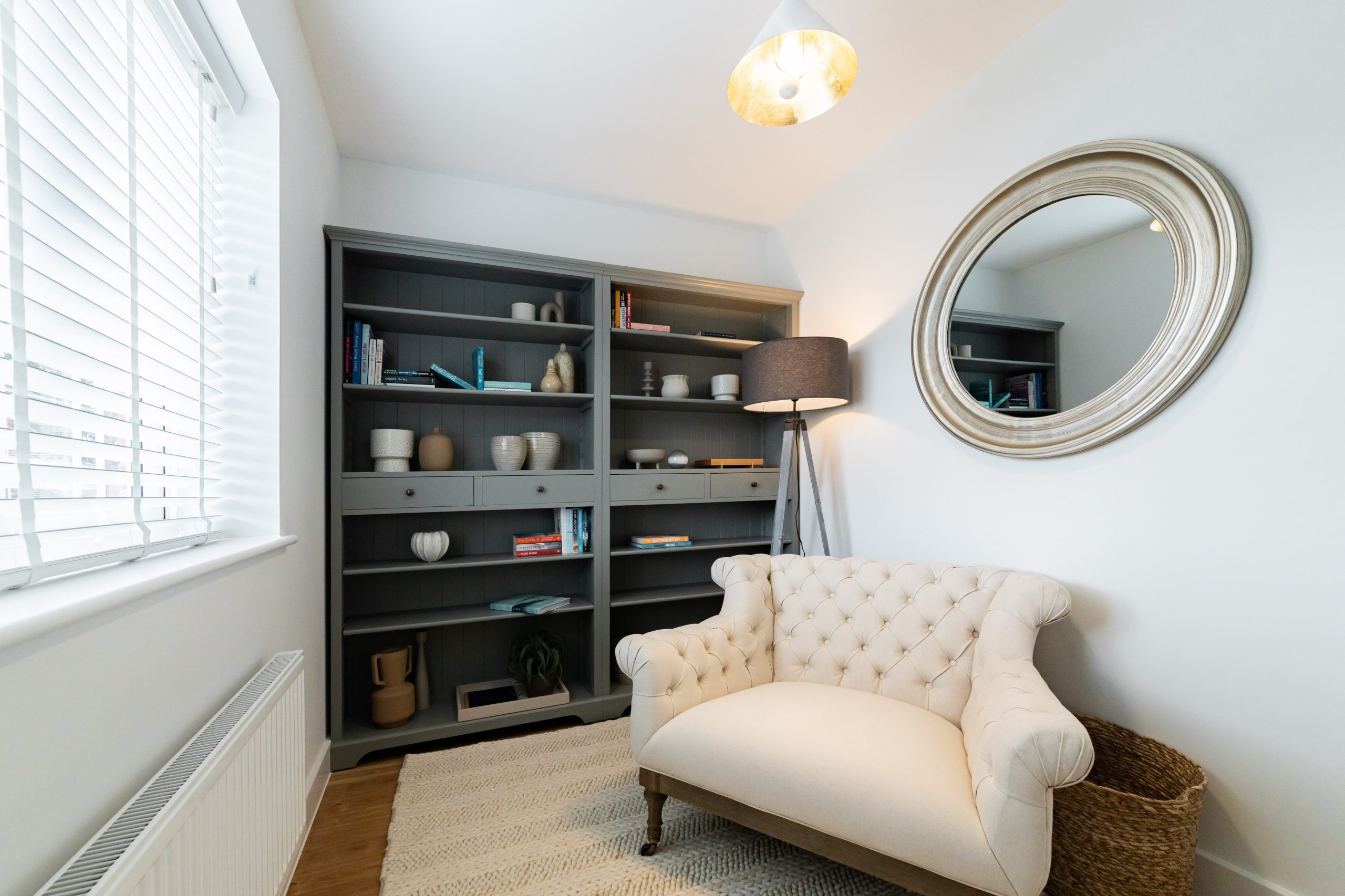 Tufted cream loveseat in a styled study with grey built-in shelves, round wall mirror, and floor lamp