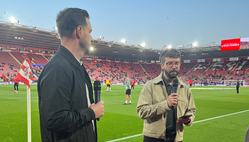 Two presenters standing pitch-side at St Mary's Stadium during a Southampton FC match, holding microphones
